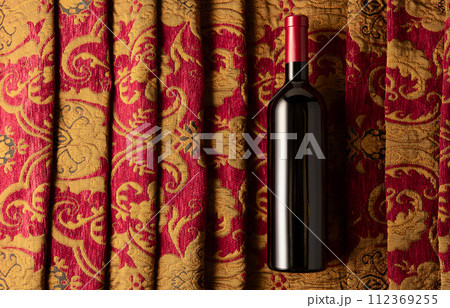 Bottle of red wine on a retro tapestry with dark red and golden floral ornament. 112369255