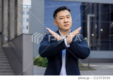 Determined male executive in smart attire gesturing prohibition with crossed arms in front of a modern building. 112372010