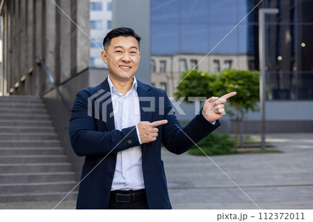 Smiling Asian businessman in a suit pointing with both hands to the side, standing on a city street with buildings in the background. Smiling Asian businessman in a suit pointing with both hands to the side, standing on a city street with buildings in the background. 112372011