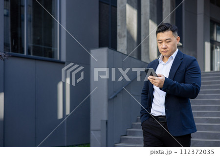 Focused Asian businessman in a suit walking outside office building, engrossed in his smartphone device. Focused Asian businessman in a suit walking outside office building, engrossed in his smartphone device. 112372035