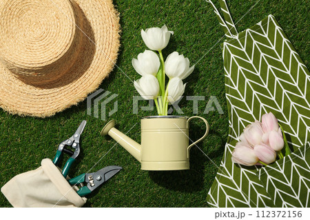 Tulips in apron and watering can, gardening tools and hat on green background, top view 112372156