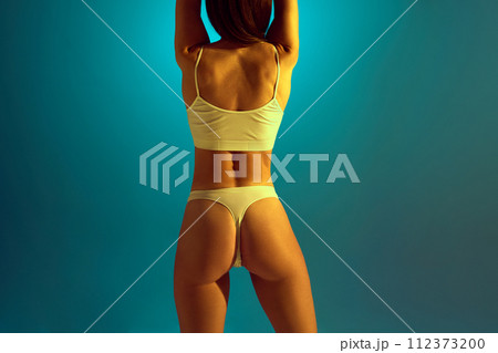 Back view of toned woman in underwear in yellow neon light against gradient studio background. Strength and vitality. 112373200