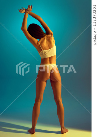 Body size portrait of beautiful, sense woman in underwear stretching back in yellow neon light against gradient studio background. 112373201