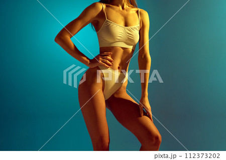 Cropped portrait of young slim woman in white underwear posing holds hands gently on leg and hip in neon light against studio background. 112373202