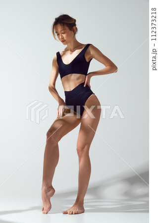 Confident, beautiful, slim woman in black swimwear posed thoughtfully against grey studio background in sun light. Protect skin from sun. Confident, beautiful, slim woman in black swimwear posed thoughtfully against grey studio background in sun light. Protect skin from sun. 112373218
