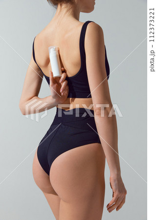 Rear view portrait of young fit woman in blue underwear holds jar of cosmetic product behind back against grey studio background. 112373221