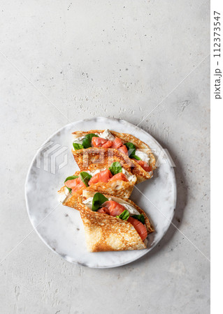 Crepes or thin pancakes with smoked salmon, soft cheese and spinach on a plate top view isolated on gray textured background. Pancake day, Maslenitsa Crepes or thin pancakes with smoked salmon, soft cheese and spinach on a plate top view isolated on gray textured background. Pancake day, Maslenitsa 112373547