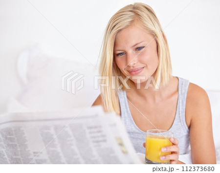 Young woman, smile and orange juice with newspaper on bed, headlines and drink for health body in wellness. Blonde model, thirsty or glass of fruit cocktail to detox, morning or vitamin c in bedroom 112373680