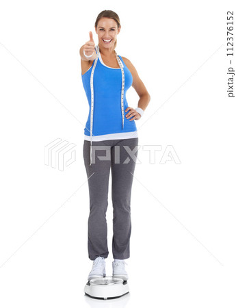 Woman thumbs up, diet scale and measure tape in studio for fitness, weight loss or health goals, like and yes. Portrait of happy model with workout results, success or feedback on a white background 112376152