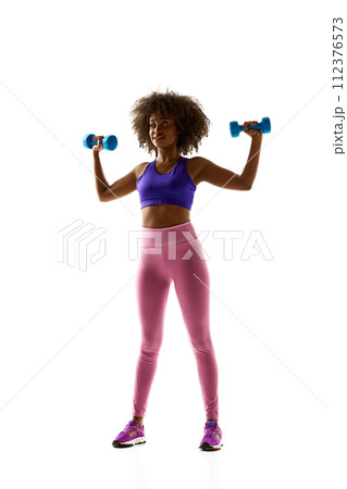 Smiling woman with dumbbells raised, wearing a purple sports bra and leggings against white studio background. Smiling woman with dumbbells raised, wearing a purple sports bra and leggings against white studio background. 112376573