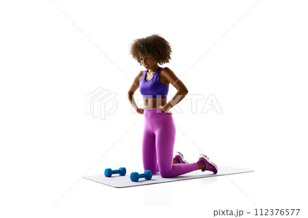 Woman in purple activewear kneeling on mat beside blue dumbbells, looking down against white studio background. Woman in purple activewear kneeling on mat beside blue dumbbells, looking down against white studio background. 112376577
