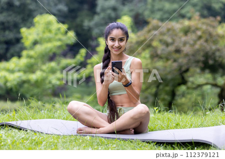 Young Indian woman in a tracksuit uses her phone while sitting on a yoga mat in a serene park, embodying fitness and relaxation. Young Indian woman in a tracksuit uses her phone while sitting on a yoga mat in a serene park, embodying fitness and relaxation. 112379121