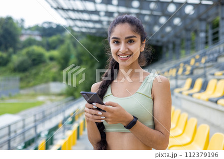 Smiling young Indian woman in a tracksuit adjusts her smartwatch during a sport and fitness routine at a daytime stadium setting. Smiling young Indian woman in a tracksuit adjusts her smartwatch during a sport and fitness routine at a daytime stadium setting. 112379196