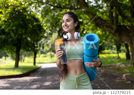 A fitness-focused Indian woman in workout attire holds a yoga mat and water bottle while enjoying a park. A fitness-focused Indian woman in workout attire holds a yoga mat and water bottle while enjoying a park. 112379215
