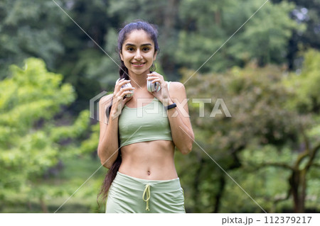 Fit Indian woman in athletic wear with headphones getting ready for a workout session in a lush park setting. 112379217