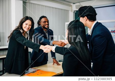 In an office, business people shake hands to celebrate an agreement. Colleagues, including marketing experts, successfully conclude a deal after a productive meeting. Teamwork 112380305