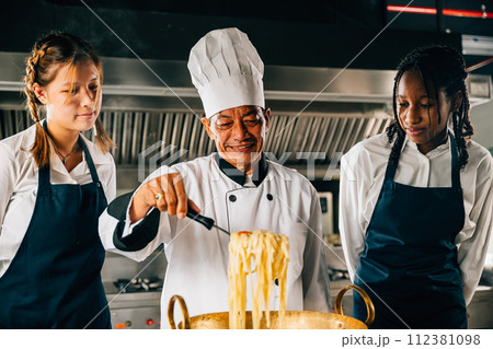 Chef guides students in kitchen. Schoolgirls cook Japanese noodle. Kids with teacher at stove. Smiling portrait of learning is modern education. Making dinner with ladle gives joy. Chef guides students in kitchen. Schoolgirls cook Japanese noodle. Kids with teacher at stove. Smiling portrait of learning is modern education. Making dinner with ladle gives joy. 112381098