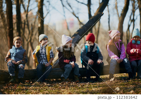 Row of kids, classmates perched on a log, clad in colorful winter wear, enjoying lesson in forest. Outdoor activities for children's development. 112381642