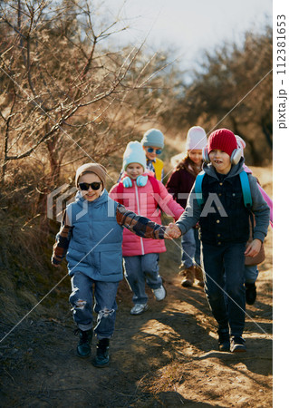 Happy, funny children holding hands wearing winter clothes and backpacks go to sunny trail. Outdoor activities for children's development. 112381653