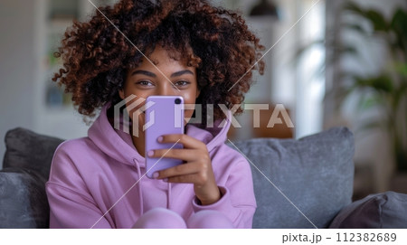 AI-generated content. Cool curly African American male teen guy model wearing pink hoodie using mobile digital apps on cellphone typing on smartphone isolated on light purple background. 112382689
