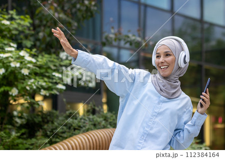 Young Muslim female student outside university campus, happy dancing outside building, woman in hijab listening to music using phone and headphones, happy smiling. Young Muslim female student outside university campus, happy dancing outside building, woman in hijab listening to music using phone and headphones, happy smiling. 112384164