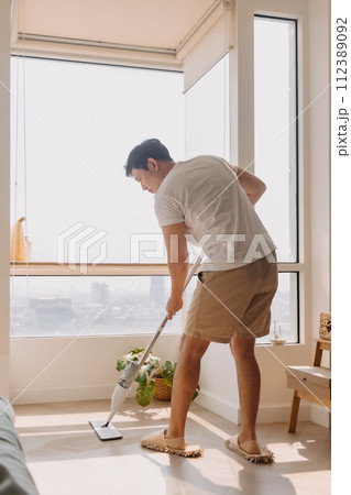 Asian Thai man using mop for cleaning floor in living room apartment, Man do household chores, housework concept. Asian Thai man using mop for cleaning floor in living room apartment, Man do household chores, housework concept. 112389092