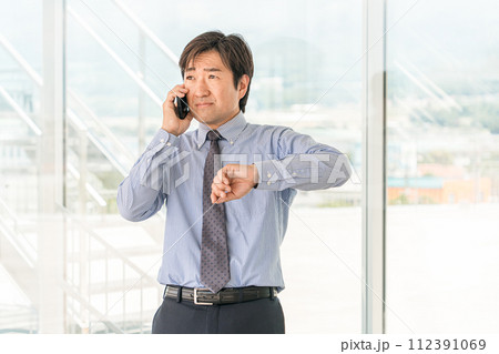 オフィスビルでスマホで通話する中年の男性アジア人ビジネスマン（遅刻・困る・トラブル・待ち合わせ） 112391069