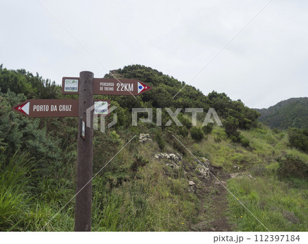 Green hills and meadow with footpath and tourist signpost pointing at Porto da Cruz in one direction and Percurso de treino at Machico in other. 112397184
