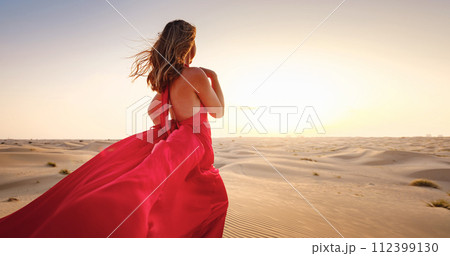 Desert adventure. Young arabian Woman in red silk dress in sands dunes of UAE desert at sunset, fantastic view. The Dubai Desert Conservation Reserve, United Arab Emirates. Desert adventure. Young arabian Woman in red silk dress in sands dunes of UAE desert at sunset, fantastic view. The Dubai Desert Conservation Reserve, United Arab Emirates. 112399130