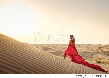Desert adventure. Young arabian Woman in red silk dress in sands dunes of UAE desert at sunset, fantastic view. The Dubai Desert Conservation Reserve, United Arab Emirates. Desert adventure. Young arabian Woman in red silk dress in sands dunes of UAE desert at sunset, fantastic view. The Dubai Desert Conservation Reserve, United Arab Emirates. 112399131