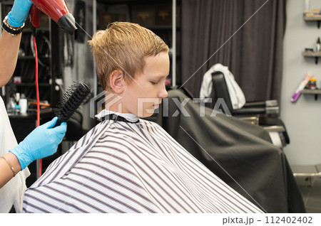 Young boy getting a haircut with hairdryer and brush at a barber shop. 112402402