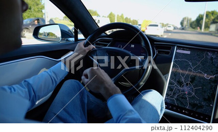 Male hand of businessman holds steering wheel while driving electric car at city. Businessperson operating electrical vehicle on urban road. Young man commuting to work at sunny day. Close up Male hand of businessman holds steering wheel while driving electric car at city. Businessperson operating electrical vehicle on urban road. Young man commuting to work at sunny day. Close up 112404290