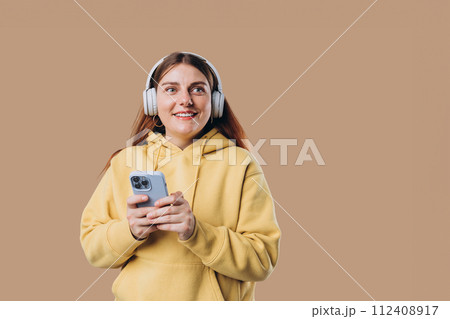 Beautiful young 30s woman wearing white wireless headphones and listening to music and using smart phone, girl isolated over colorful background wall Beautiful young 30s woman wearing white wireless headphones and listening to music and using smart phone, girl isolated over colorful background wall 112408917