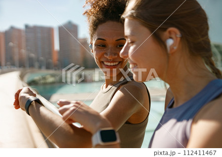 Smiling woman with female friend looking on smartwatch before exercising standing outdoors 112411467