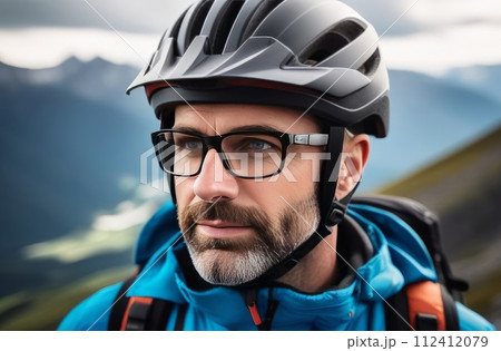 Man wearing helmet and glasses stands confidently before towering mountain backdrop ready for adventure and exploration. He may be gearing up for bicycle ride or some other outdoor activity. 112412079
