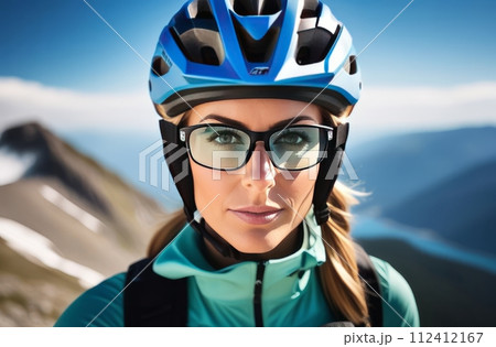 Woman wearing helmet and glasses stands confidently before towering mountain backdrop ready for adventure and exploration.She may be gearing up for bicycle ride or some other outdoor activity. 112412167