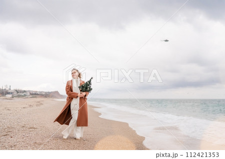 Blond woman Christmas tree sea. Christmas portrait of a happy woman walking along the beach and holding a Christmas tree in her hands. She is wearing a brown coat and a white suit. 112421353