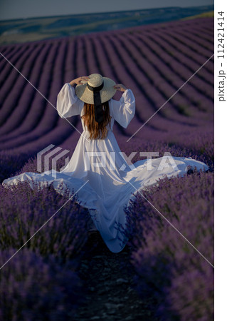 Back view woman lavender sunset. Happy woman in white dress holds lavender bouquet. Aromatherapy concept, lavender oil, photo session in lavender Back view woman lavender sunset. Happy woman in white dress holds lavender bouquet. Aromatherapy concept, lavender oil, photo session in lavender 112421414