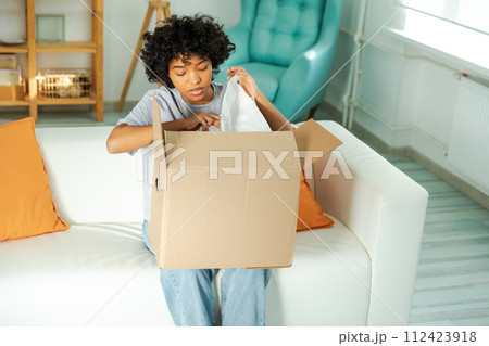 African girl unpacking delivery looking in box. Happy woman opening carton box. Female getting parcel looking at delivered goods items. Satisfied client positive feedback quick express deliver 112423918