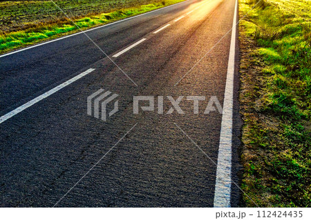 An open road with white lines, bordered by sunlit grassy fields. 112424435