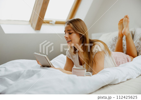 Young smiling woman reading notes in note pad while sitting on bed at home  112425625