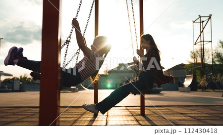 Happy female children silhouette swinging swing at summer sunset sunrise sky outdoor sport playground. Girl kids adolescent having fun flying enjoy leisure activity childhood recreation joyful playing 112430418