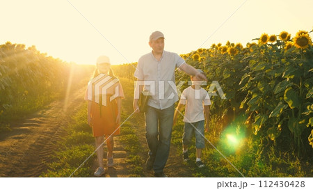 Family father and kids walking at sunflower field pointing on yellow flowers sunset sun light. Man with daughter and son going surrounded by agriculture harvest floral plant meadow natural sunlight Family father and kids walking at sunflower field pointing on yellow flowers sunset sun light. Man with daughter and son going surrounded by agriculture harvest floral plant meadow natural sunlight 112430428