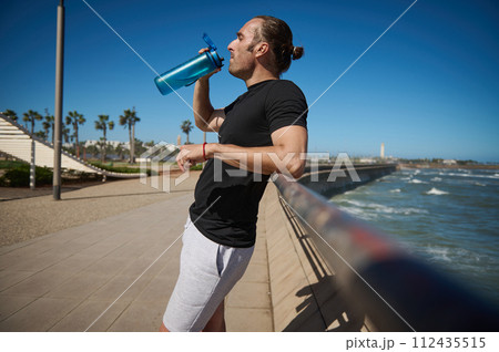 Determined athlete man drinking water from sports bottle, resting after early morning jog at Atlantic ocean promenade Determined athlete man drinking water from sports bottle, resting after early morning jog at Atlantic ocean promenade 112435515