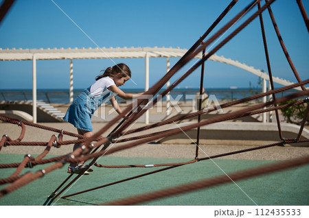 Adorable little girl climbing on a climbing net at the city playground on the embankment. 112435533