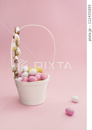 White Easter basket with chocolate multicolored eggs and willow branch on pastel pink background. Easter composition. Card with copy space for text White Easter basket with chocolate multicolored eggs and willow branch on pastel pink background. Easter composition. Card with copy space for text 112435895
