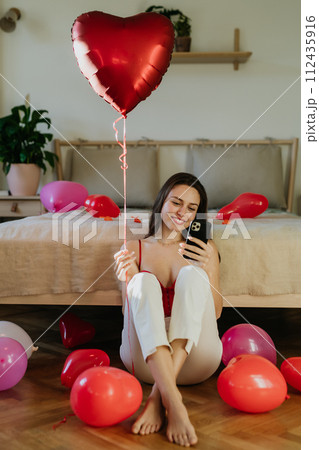 Beautiful woman sitting by bed in the middle of baloons, scrolling on smartphone. Concept of Valentine's Day, love and romance. 112435916