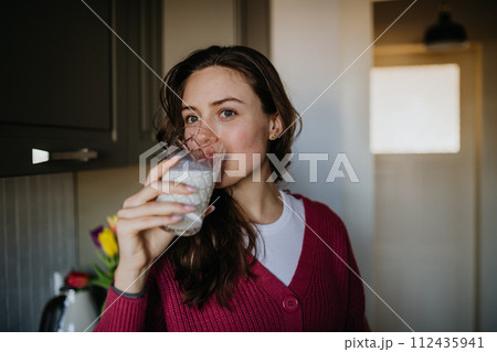 Beautiful woman drinking glass of plant-based milk in the kitchen. 112435941