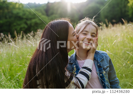 Beautiful mother with daughter, embracing, kissing on cheek, sitting in the grass at meadow. Concept of Mother's Day and maternal love. 112436280