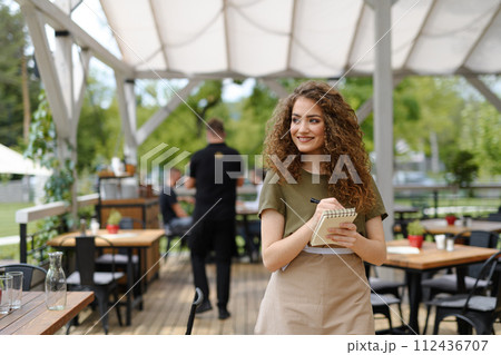 Portrait of a beautiful waitress writing down an order in paper notebook with pen. Server standing on restaurant terrace in an apron. Portrait of a beautiful waitress writing down an order in paper notebook with pen. Server standing on restaurant terrace in an apron. 112436707
